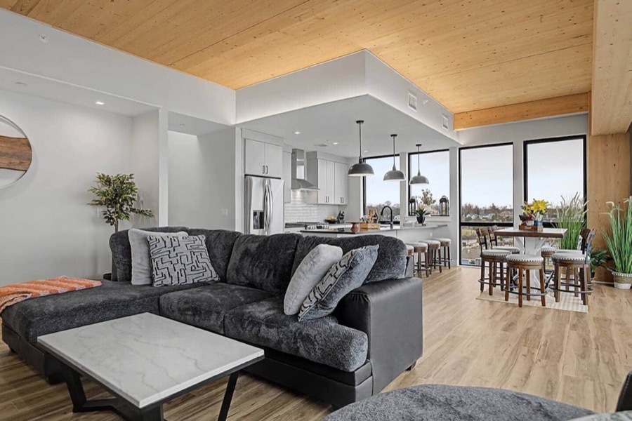 The 601-wing great room — charcoal sectional under the mass-timber ceiling, with the white island kitchen and dining table beyond