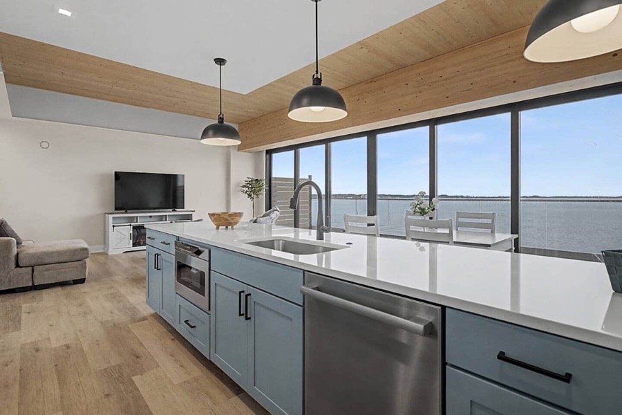 Open kitchen and living area of the Above the Marina penthouse with a long quartz island, timber ceiling, and floor-to-ceiling windows framing Muskegon Lake