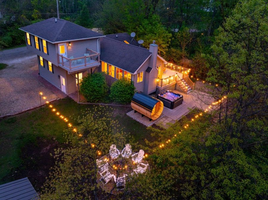 Aerial dusk view of Bell Cottage: the lit deck and balcony, barrel sauna and hot tub patio, and a lantern-lined path winding to the fire-pit circle