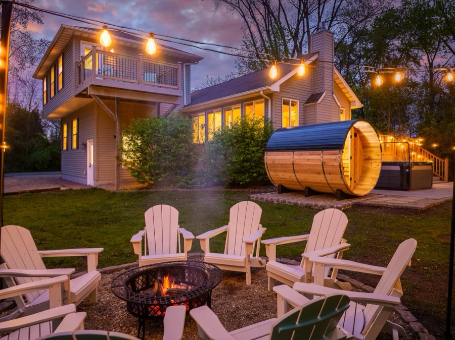 Bell Cottage's backyard at dusk — Adirondack chairs circling a crackling fire pit, with the cedar barrel sauna, hot tub, and string-lit cottage behind