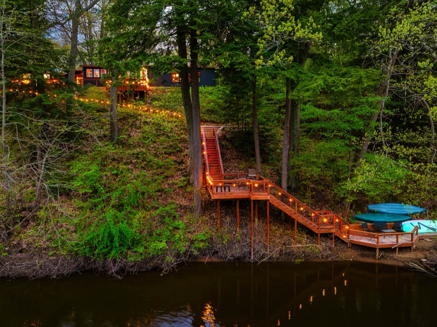 String-lit wooden staircase winding down a wooded bank at dusk to a riverside deck with kayaks on the Kalamazoo River