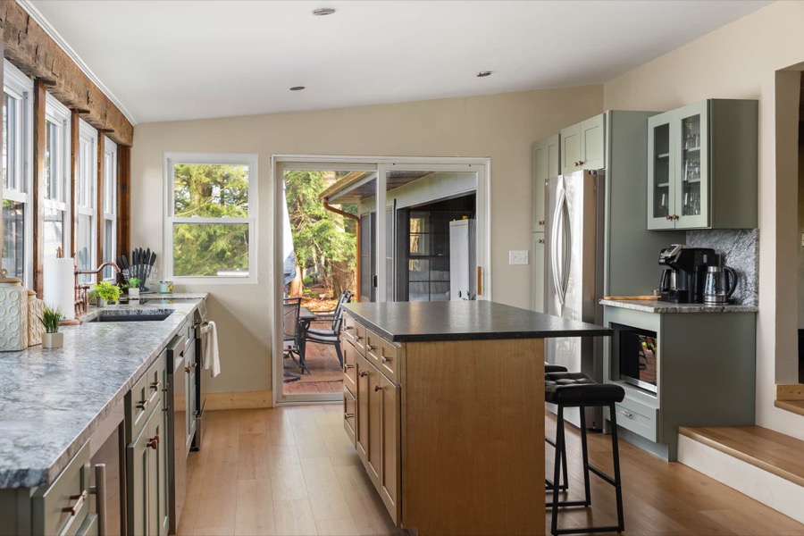 The redesigned chef's kitchen: sage-green cabinetry, granite counters, a big island with seating, and sliders opening to the deck