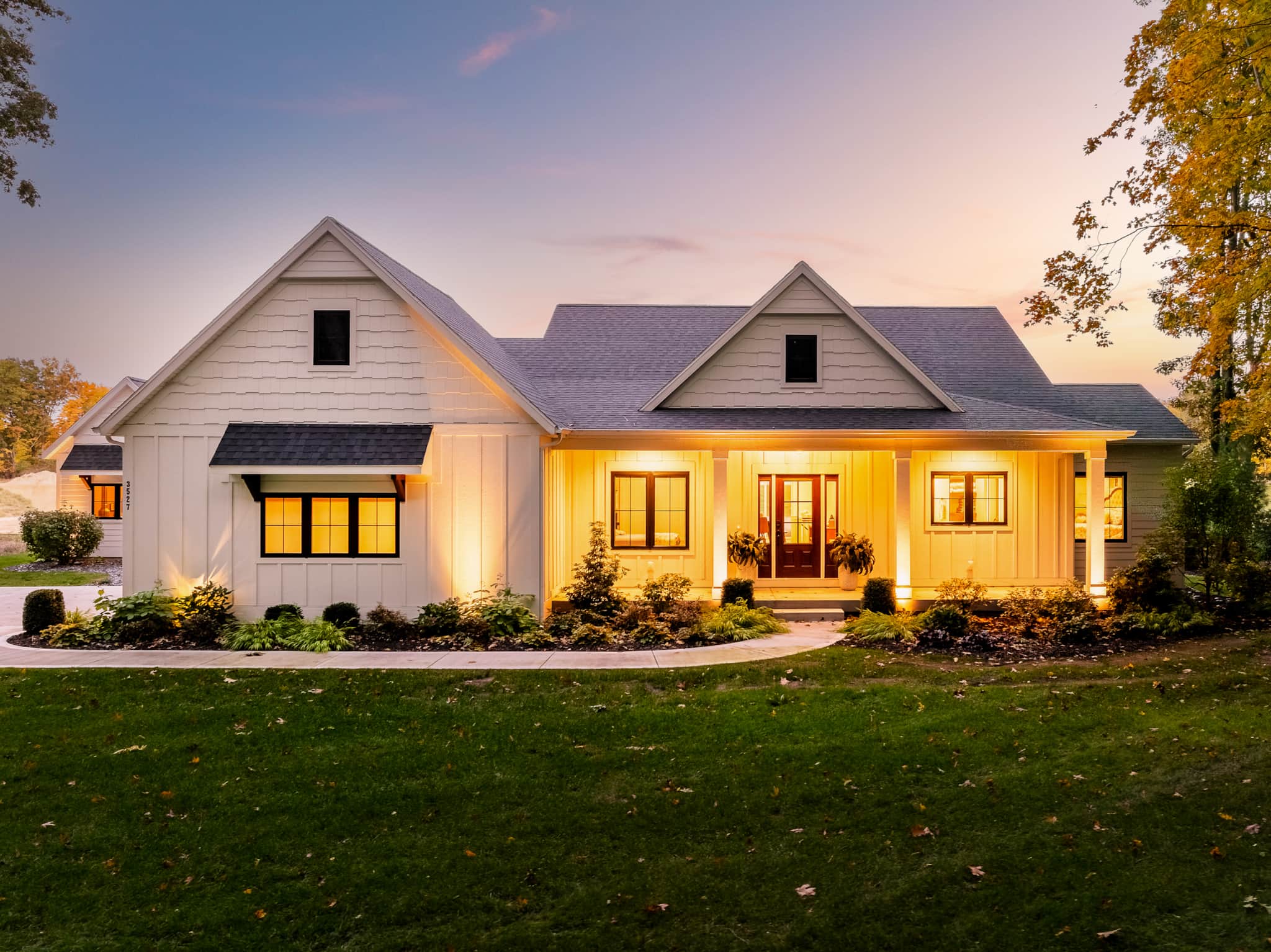Modern farmhouse-style vacation home glowing warmly at dusk, its white board-and-batten facade lit against a violet evening sky in Saugatuck, Michigan