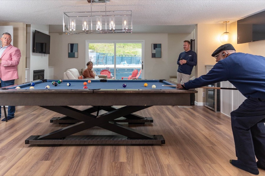 A family game of pool in the game room, with the patio doors framing the backyard pool and red Adirondack chairs